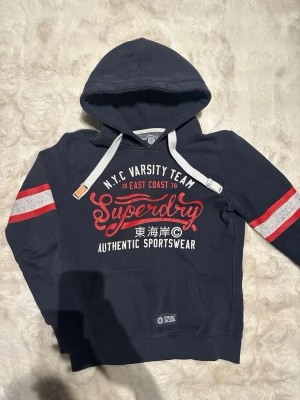  Superdry hoodie - Mörkblå hoodie från Superdry med vita och röda detaljer. Har luva med vita snören och röda samt vita ränder på ärmarna. Storlek XS. OBS på bild 4 finns ett jättelitet hål men det märks inte av, hålet är typ 2 mm. Skick 8/10. Perfekt för en sportig och avslappnad stil. Endast 450 priset är ej hugget i sten!
