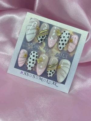 Bijou (XS);Medium Almond - Snygga press-on naglar i mandelform från VonNija Nails. Mix av ljusrosa, nude och vita nyanser med svarta prickar, vita linjer och guldfärgade dekorationer. Några naglar har små guldfjärilar och kedjedetaljer för extra bling. Perfekt för dig som vill sticka ut.