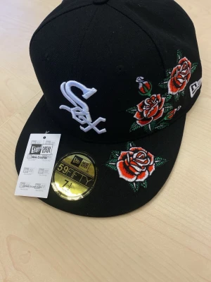 Svart New Era keps med rosbrodyr SOX - Snygg svart keps från New Era, modell 59FIFTY, med broderade röda rosor och vita detaljer. Framsidan har ett vitt Sox-logo broderi och kepsen har en platt skärm. Perfekt för dig som vill sticka ut med en unik accessoar. fitted cap