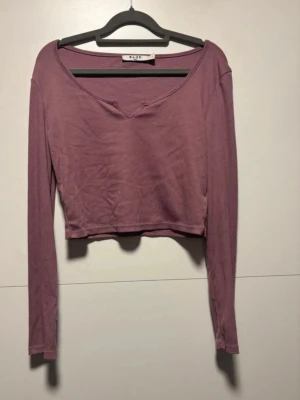 Lila långärmad crop top från NA-KD - Trendig lila långärmad crop top från NA-KD med djup v-ringning och mjukt, stretchigt material. Perfekt att styla med höga jeans eller kjol för en avslappnad look. Storlek S. 