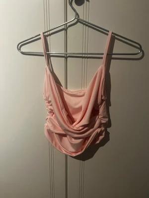 Ljusrosa draperad linnetopp - Trendig ljusrosa linnetopp med draperad front och smala axelband. Toppen har en croppad passform och är tillverkad i ett mjukt, stretchigt syntetmaterial som ger en snygg siluett. Perfekt att styla med höga jeans eller kjol.