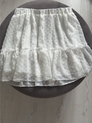 Vit volangkjol med prickigt mönster - Söt vit kortkjol med volang och prickigt mönster. Kjolen har resår i midjan och är gjord i ett lätt, transparent material med dubbla lager för extra volym. Perfekt för en trendig och feminin look.