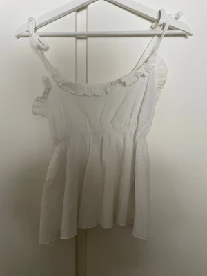 Vit linnetopp med volang från 157 - Supersöt vit linnetopp från 157 med volangdetaljer längs ringningen och knytbara axelband. Toppen är ärmlös och har en luftig, flowy passform som passar perfekt till sommaren. Materialet känns lätt och skönt mot huden.