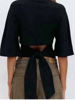 Svart croppad topp med knyt - Trendig svart croppad topp med halvlånga ärmar och snygg knytning i ryggen. Toppen har en öppen detalj baktill och är perfekt för dig som gillar en stilren men ändå edgy look. Passformen är normal och materialet känns mjukt mot huden.
