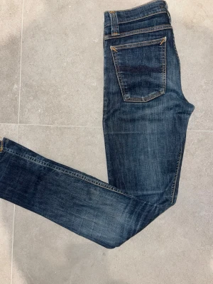 Nudie Jeans - Säljer ett par blå smala jeans från Nudie Jeans med klassisk femficksdesign och snygga slitningar. Jeansen har orange sömmar och en patch med Nudie-loggan bak i midjan. Perfekta för dig som gillar en avslappnad men ändå stilren look.