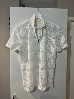 Vit, transparent spetsblus med korta ärmar - Säljer en vit blus i spets med blommönster och korta ärmar. Blusen har en liten krage och v-ringning med dekorativ dragkedja framtill. Perfekt för dig som gillar detaljer och en ljus, fräsch look.