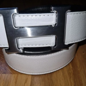 Vitt skärp med stor silverspänne - Snyggt vitt skärp med ett stort H-format spänne i silverfärg. Skärpet har en stilren och modern look med slät yta och bred design. Perfekt accessoar för att lyfta din outfit.