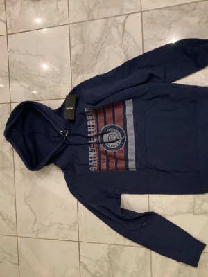 Mörkblå saint Laurent hoodie - Snygg mörkblå hoodie från Saint Urbain med huva och dragsko. Framsidan har ett stort tryck i vitt och rött med text och emblem. Tillverkad i mjukt bomullsmaterial och har en klassisk känguruficka framtill. Perfekt för en avslappnad streetwear-look.