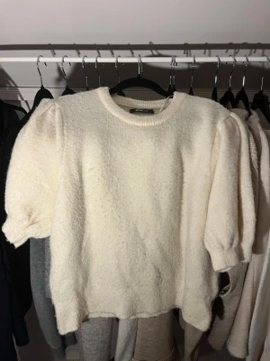 Krämvit stickad puffärmströja - Mysig krämvit stickad tröja från GINA TRICOT med korta puffärmar och rund halsringning. Tröjan har en mjuk, fluffig struktur och ribbade kanter vid hals och ärmslut. Perfekt för dig som gillar en söt och trendig look.