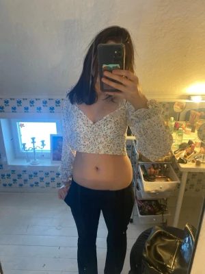 Blommig croppad omlottopp - Säljer en vit croppad omlottopp med tyg som silke, blommigt mönster och långa ärmar. Toppen har dragkedja på ryggen, vilket ger en snygg och trendig look. Perfekt för dig som gillar magtröjor och vill ha något som sticker ut.