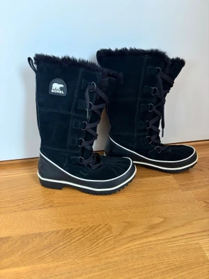 Svarta vinterstövlar från Sorel - Svarta höga stövlar från Sorel med snörning framtill och mjukt foder upptill. Tillverkade i mocka och textil med robust sula och vit rand längs kanten. Perfekta för kalla dagar och har en klassisk outdoor-look. Små 37:or.