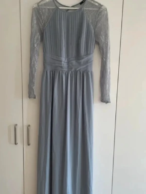 Ljusblå långklänning med spetsärmar - Snygg ljusblå långklänning med plisserad överdel och transparenta spetsärmar. Klänningen har markerad midja och rund halsringning. Perfekt för dig som vill ha en elegant look med romantiska detaljer.