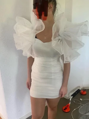 Coolasteeee studentklänningen😁😁 - Supersnygg vit kortklänning med stora, dramatiska volangärmar och smala axelband. Klänningen har en figurnära passform och är perfekt för dig som vill sticka ut. Materialet ser ut att vara lätt och luftigt, troligen polyester eller syntet.