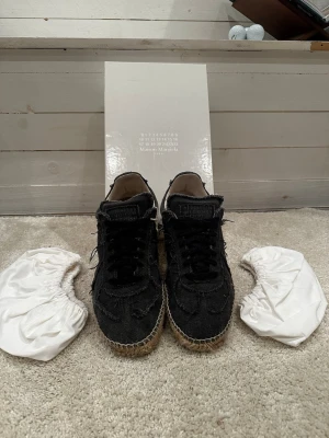  Maison Margiela gats - Unika svarta sneakers från Maison Margiela med råa kanter och snörning. Ovandelen är i textil med en cool, fransad finish och sulan har ett espadrille-inspirerat flätat mönster. Skorna har rund tå och klassisk sneakerform. Perfekta för dig som gillar statement-plagg.
