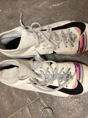 Nike vita fotbollsskor med färgdetaljer - Säljer ett par vita Nike fotbollsskor med högt skaft och gråa snören. Skorna har en stor svart swoosh på sidan och färgglada ränder vid tån. Perfekta för dig som vill sticka ut på planen.