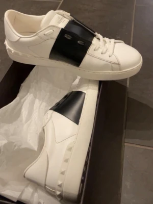 Valentino Garavani vita sneakers - Snygga vita sneakers från Valentino Garavani med bred svart läderdetalj över snörningen och ikoniska nitar på hälen. Skorna har en stilren och modern design i premiumläder och passar perfekt till streetwear eller en clean look. Skon har fått en liten miss färgning från jeans men de går att få bort.