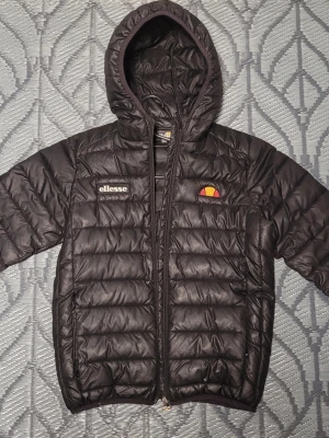 Svart dunjacka från Ellesse - Snygg svart dunjacka från Ellesse, lite tunnare med huva och dragkedja framtill. Jackan har Ellesse-logga på bröstet och quiltad design som ger en sportig look. Perfekt för kyliga dagar och enkel att matcha med olika outfits.