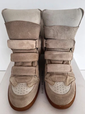 Beige höga sneakers Isabel Marant - Snygga höga sneakers från Isabel Marant i beige mocka och vitt läder. Skorna har flera kardborreband och en chunky brun sula. Perfekta för dig som gillar streetstyle och vill ha något unikt till din outfit.