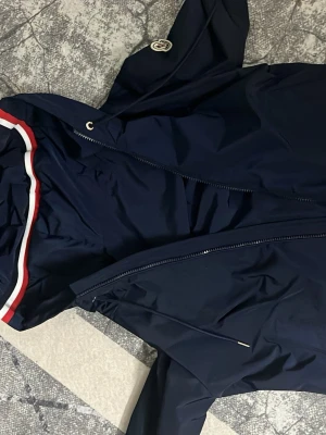 Mörkblå vindjacka från Moncler - Snygg mörkblå vindjacka från Moncler med dragkedja, huva och vita samt röda detaljer längs huvkanten. Jackan har en tydlig Moncler-logga på bröstet och är tillverkad i ett lätt, vattenavvisande material. Perfekt för dig som vill ha en stilren och sportig look.