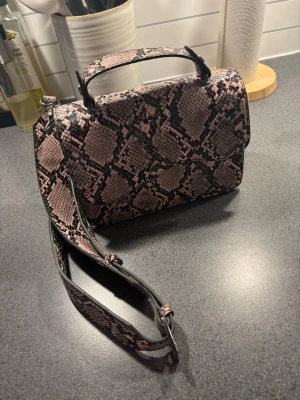 Rosa ormmönstrad handväska - Säljer en snygg handväska i rosa och svart ormmönstrat konstläder. Väskan har både handtag och justerbar axelrem, perfekt för att bära på olika sätt. Stilren och trendig design med lock och magnetknäppning. Perfekt accessoar för att lyfta din outfit.