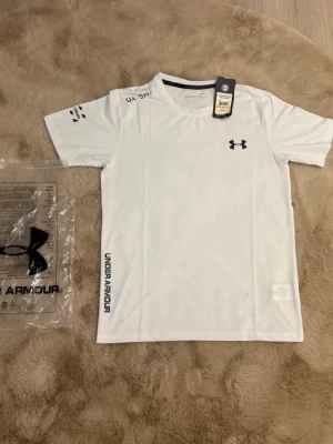 Vit träningströja från Under Armour - Vit kortärmad träningströja från Under Armour med svart logga på bröstet och tryck på ärmen samt sidan. Tillverkad i ett lätt och snabbtorkande material som passar perfekt för träning och sportaktiviteter.