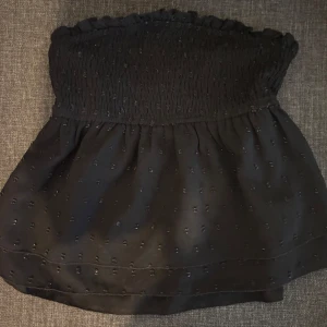 Svart topp - Säljer en svart bandeau tubtopp med små diskreta prickar och smock upptill. Toppen är axelbandslös och har en lätt volangkant upptill för en söt touch. 
