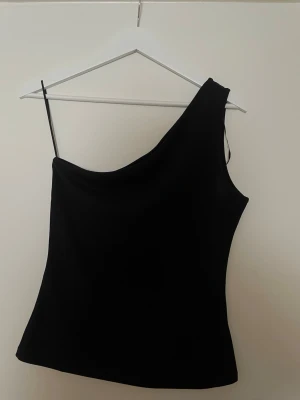 Svart oneshoulder topp från H&M - Snygg svart oneshoulder topp från H&M med en axelbandslös sida och ett smalt axelband på andra sidan. Enkel och stilren design som passar perfekt till många olika looks. Tillverkad i mjukt och stretchigt material. Aldrig använd