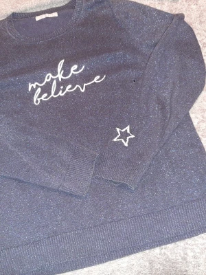 Mörkblå stickad tröja med glitter - Mörkblå stickad tröja med glittrigt material och vit text 'make believe' framtill. Tröjan har en broderad vit stjärna på ena ärmen, rund halsringning och ribbade muddar vid ärmslut och nederkant. Perfekt för dig som gillar lite extra detaljer.