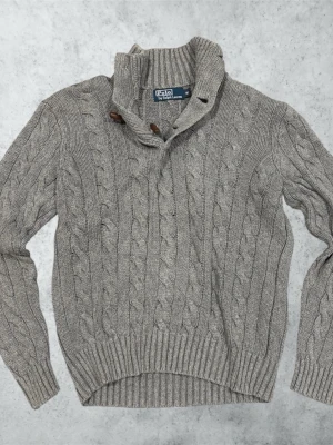 Grå kabelstickad tröja Polo Ralph Lauren - Säljer en grå kabelstickad tröja från Polo Ralph Lauren med hög krage och träknappar. Tröjan har ribbade muddar och nederkant, samt ett klassiskt flätmönster över hela plagget. Perfekt för dig som gillar stilren och tidlös design. Mått: Bredd; 46 cm. Längd; 62 cm. Ärm; 60 cm