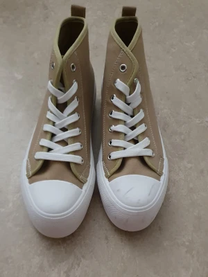 Sneakers - Säljer ett par beige höga sneakers med vita gummisulor och tåhättor. Skorna har vita skosnören och metallhål för snörningen. Tillverkade i canvasmaterial och har en stilren, klassisk design som passar till det mesta. Några fläckar.