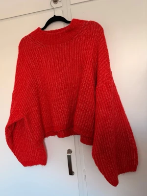 Röd oversized stickad tröja - Säljer en röd oversized stickad tröja med breda ärmar och ribbad struktur. Underbar röd-orangefärg i verkligheten. Tröjan har en hög, rund halsringning och är croppad i modellen. Perfekt för dig som gillar en avslappnad och trendig stil. Obs. hund finns i hemmet 
