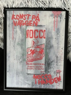 Nocco skumnisse tavla - Unik tavla med motiv av NOCCO skumnisse, gjort i street art-stil med röd och grå färg. Texten 'KONST PÅ VÄGGEN' och 'NOCCO I HANDEN' är sprayade på duken. Svart ram ingår. Perfekt för dig som gillar popkultur och energidryck.