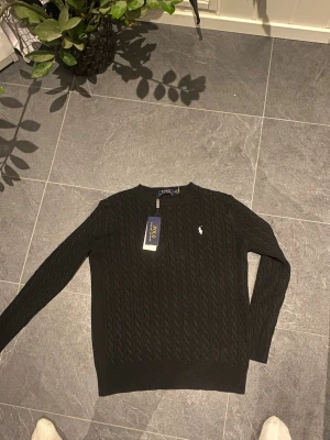 Svart kabelstickad tröja från Polo Ralph Lauren - Svart kabelstickad tröja från Polo Ralph Lauren med klassisk rund hals och diskret broderad logotyp på bröstet. Tröjan har långa ärmar och ribbade muddar vid ärmslut och nederkant. Perfekt för dig som gillar stilrena och tidlösa plagg.