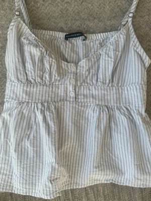 Randig linnetopp från Brandy Melville - Säljer en söt vit och ljusblå randig linnetopp från Brandy Melville. Toppen har smala axelband, knappar framtill och en lätt rynkad midja som ger en avslappnad look. Perfekt för varma dagar och enkel att styla med jeans eller kjol.