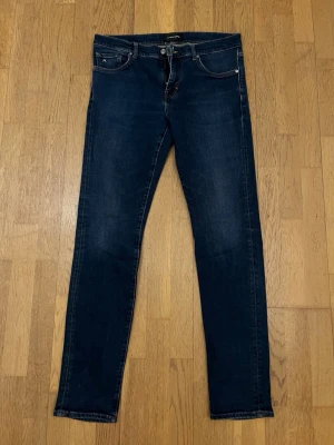 Mörkblå skinny jeans J.Lindeberg - 34/34 - Snygga mörkblå skinny jeans från J.Lindeberg, modell Jay. Klassisk femficksdesign med silverfärgade nitar och knapp. Jeansen har en smal passform och är tillverkade i ett stretchigt denimtyg för extra komfort. Perfekta för en trendig och avslappnad look.
