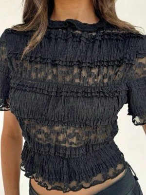 Svart transparent volangblus - svart blus med transparent tyg och volangdetaljer. Kortärmad modell med hög hals. Vet inte vart tröjan är ifrån för jag fick den av min kusin. 