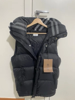 Svart dunväst från Burberry - Stilren svart dunväst från Burberry med stor, avtagbar huva och grått mönstrat foder. Västen har dragkedja och knappar framtill, samt två praktiska fickor med dragkedja. Perfekt för lager-på-lager och streetwear-vibe.