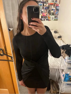 Svart omlottklänning med lång ärm - Snygg svart kortklänning med omlottdetalj framtill och långa ärmar. Klänningen har en tight passform och är tillverkad i ett mjukt syntetmaterial som sitter bekvämt. Perfekt för dig som gillar stilrena och moderna plagg.