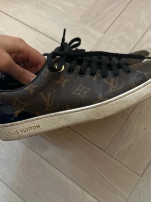Louis Vuitton monogram sneakers - Snygga sneakers från Louis Vuitton med klassiskt brunt monogrammönster, svarta skosnören och vita sulor. Skorna har guldfärgade detaljer vid snörningen och är tillverkade i läder med canvas-överdrag. Perfekta för dig som gillar lyxig streetstyle.