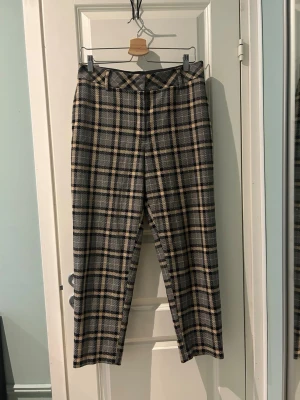Rutiga ankellånga byxor i beige och svart - Snygga kostymbyxor med rutigt mönster i beige, svart och grått. Byxorna är ankellånga med bälteshällor och klassisk normalhög midja. Perfekta för dig som vill ha en stilren men trendig look! Storlek 42