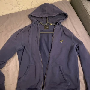 Mörkblå hoodie från Lyle & Scott - Säljer en snygg mörkblå hoodie från Lyle & Scott i bra skick. Den har en dragkedja framtill och en klassisk gul logga på bröstet. Perfekt för höst och vår, med långa ärmar och en skön huva. Passar både till vardags och chillkvällar. 