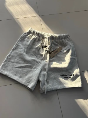 Grå Essentials shorts med snörning - Snygga grå shorts från Fear of God Essentials med broderad logga på benet. Modellen har elastisk midja med snörning och är gjorda i mjuk bomull för maximal komfort. Perfekta för en avslappnad och trendig look.