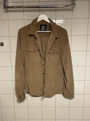 Beige overshirt i manchester från Dickies - Snygg beige overshirt från Dickies i mjuk manchester. Klassisk krage, två bröstfickor med lock och knappar framtill. Perfekt lager-på-lager-plagg med avslappnad passform och långa ärmar. Enkel och stilren design som funkar till många outfits.