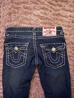 True Religion jeans med kontrastsömmar - Säljer ett par mörkblå True Religion jeans med ikoniska vita kontrastsömmar och stora bakfickor med knappdetaljer. Modellen har låg midja och raka ben, klassisk logopatch baktill och coola metalldetaljer. Tillverkade i USA av slitstarkt denim. Dom e väldigt lowrise, passar xs eller xxs 