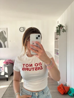 Vit cropad t-shirt med röd text - Vit, tight och cropad t-shirt med trycket 'DO NOT DISTURB' i stora röda bokstäver på bröstet. Perfekt för dig som gillar statement-plagg och enkel stil. Rund halsringning och korta ärmar.