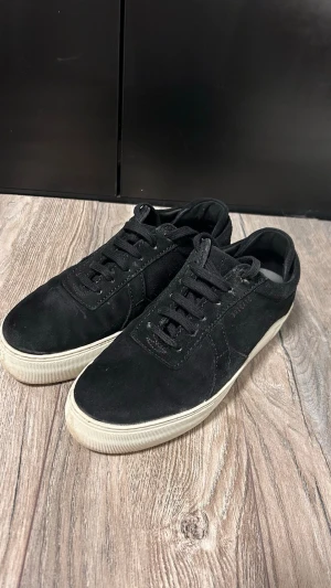 Svarta sneakers i mocka - Stilrena svarta sneakers i mocka med klassisk låg profil och vita sulor. Skorna har svarta skosnören och diskreta sömmar längs sidorna. Perfekta för en clean och enkel look.