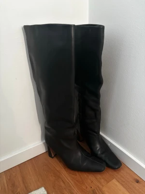 Svarta knähöga boots med klack - Säljer ett par svarta knähöga stövlar i slätt skinnimitation med spetsig tå och bred klack. Stövlarna har en stilren och minimalistisk design som passar till många olika outfits. Perfekta för dig som vill ha ett par klassiska boots med lite höjd.