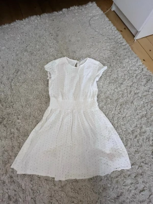 Vit kort klänning med broderade hål - Söt vit kortklänning med broderade hål och resår i midjan. Klänningen har korta ärmar och rund halsringning. Perfekt för varma dagar och ger en romantisk vibe med sina fina detaljer.