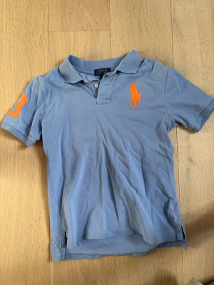 Blå pikétröja från Polo Ralph Lauren - Säljer en ljusblå pikétröja från Polo Ralph Lauren med klassisk krage, korta ärmar och knappar vid halsen. Tröjan har en stor orange broderad logga på bröstet och siffran 3 på ärmen. Tillverkad i bomull för en skön och avslappnad stil.