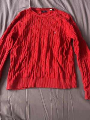 Röd kabelstickad tröja från Gant - Säljer en klassisk röd kabelstickad tröja från Gant med rund halsringning och diskret logga broderad på bröstet. Tröjan har långa ärmar och ribbade muddar vid ärmslut och nederkant. Perfekt för dig som gillar tidlös och stilren design.